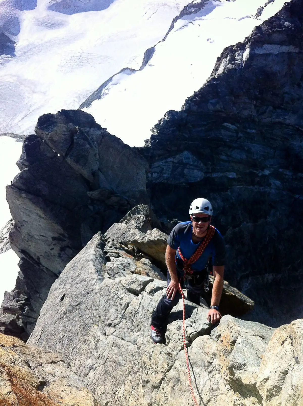 Man ascending a rocky structure with help of a rope, wearing sunglasses and a helmet. In the background the mountains are snowy.